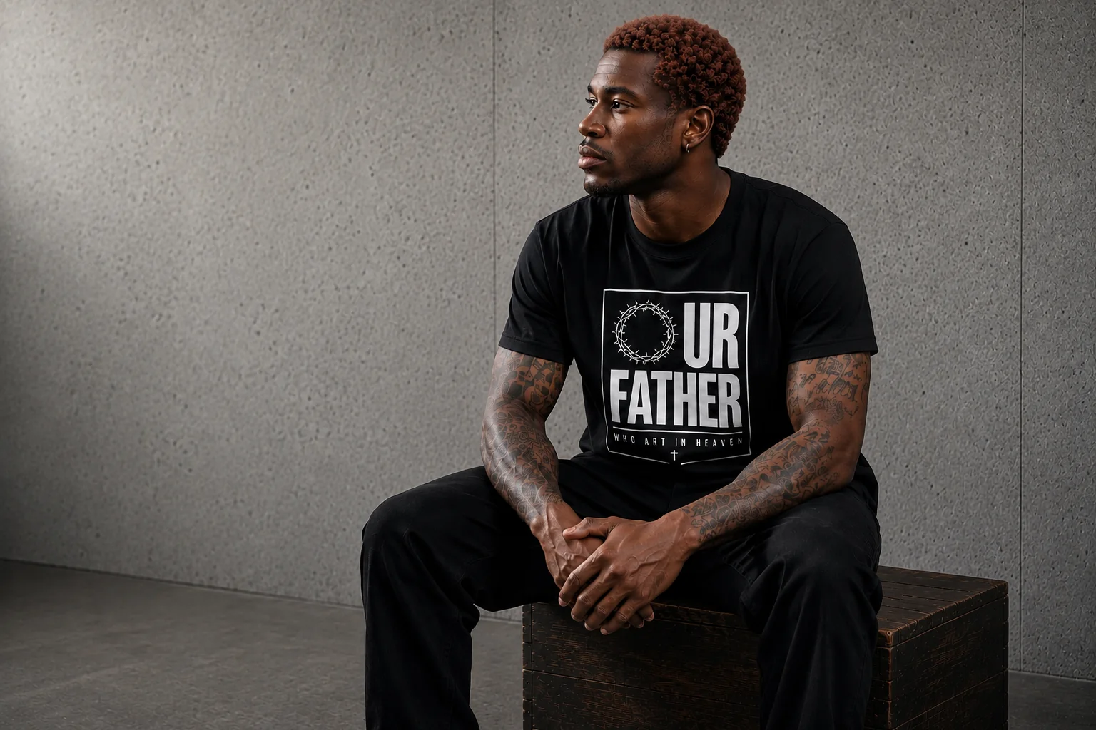 Man seated against a concrete wall wearing the Our Father tee